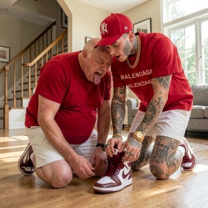 Dos hombres arrodillados sobre un suelo de madera en un salón luminoso parecen ajustar o atar los cordones de una zapatilla deportiva roja y blanca. Ambos visten camisetas rojas y pantalones cortos claros; el hombre mayor, con la lengua fuera y expresión concentrada o juguetona, lleva zapatillas granates, mientras el hombre más joven luce una gorra roja de los Yankees, camiseta con el texto Balenciaga, reloj dorado y numerosos tatuajes en brazos, cuello y piernas. La escena transcurre en una casa moderna con escalera de madera al fondo, grandes ventanas, sofá gris y luz natural, creando un ambiente doméstico informal y cercano.