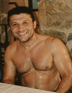 A smiling man appears seated next to a table with a white tablecloth, shirtless and with shiny skin, as if damp from sweat or heat. He has short dark hair, stubble, a relaxed expression, and a direct gaze at the camera. His chest and abdomen show abundant hair, with robust shoulders and arms visible in the foreground. The setting seems rustic and indoors, with an irregular stone wall in the background and dark wooden elements suggesting a restaurant, wine cellar, or country house. The warm lighting highlights the texture of the skin, the male torso, and the traditional stone surroundings.