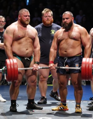 Varios hombres extremadamente musculosos y barbudos en una competición de halterofilia. Se muestra con una pequeña camiseta que deja ver sus enormes hombros en su pecho, lleva un pequeño pantalón corto que marca paquete mientras levanta una enorme barra con pesas de trescientos kilos