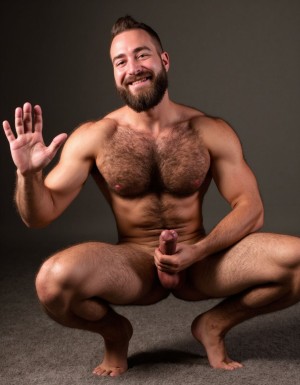Hombre con mucho vello corporal musculoso totalmente desnudo agachado con pene erecto y saluda con la mano izquierda y con la mano derecha rodea su pene erecto. Sonríe