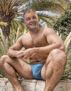 A smiling man squats outdoors, bare-chested and wearing light blue brief-style swim trunks, in a tropical setting with palm trees, ornamental plants, and tall grasses in the background. His muscular, tanned body appears slightly wet, with visible hair on his chest, abdomen, arms, and legs, reinforcing a summer, beach, or pool aesthetic. He is perched on a stone wall, with his legs open and his arms resting on his knees, looking directly at the camera with a relaxed and cheerful expression under the intense sunlight.