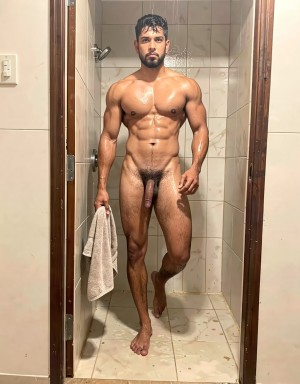 Naked man with a very muscular build posing standing under the shower, with a wet and shiny body, defined abs, defined chest, strong arms, and athletic legs. He is facing the camera inside a narrow bathroom with light tiles, a dark wooden screen or frame, and a wet tiled floor; he holds a beige towel in one hand while maintaining a firm and direct posture. He has wet dark hair, a trimmed beard, and a serious expression. Body hair is visible on his chest, abdomen, pubic area, and legs, and the penis is exposed frontally in the center of the composition.