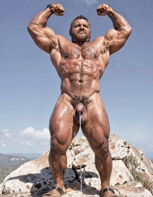 A hyper-masculine ancient Zapotec warrior named Cocijo, 26 years old, standing perfectly still under the brutal midday sun on the rocky summit of Cerro La Cantera in San Pablo Huitzo, Oaxaca, 600 AD. Completely naked except for worn leather strip sandals. Square jaw clenched, thick veiny neck, bronze skin drenched in sweat. Extremely powerful and fully flexed physique: massive square pectorals tensed to the maximum, thick dark wet chest hair matted against the hard muscle, large erect nipples. Deeply carved eight-pack abs glistening with sweat, thick black treasure trail running down to a dense dark pubic bush. Enormous arms raised and flexed hard overhead, biceps bulging like cannonballs with thick veins, triceps perfectly defined and tensed like horseshoes. Thick hairy thighs locked in full tension. His massive 35cm cock is rock-hard and throbbing violently, held tightly at the very base by Gayon Von Neumann’s strong hand, the swollen head continuously leaking thick white precum that runs down the shaft and over the fingers. A thick black leather cock ring is locked brutally tight at the root, behind his heavy balls, making the entire package look painfully swollen, darker and pulsing non-stop. Cocijo stares straight up with total obedience, every muscle in his body still tensed and displayed for inspection. Raw, dominant, extremely virile ancient masculine atmosphere, dramatic sunlight casting strong shadows on every ridge of muscle, hyper-realistic and extremely detailed.