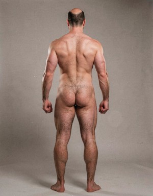Mike Morris, a 32-year-old extremely lean Caucasian male at only 8% body fat, stands completely naked in a strict military attention pose seen from directly behind in a full posterior view. His back is perfectly straight, shoulders pulled back, arms rigid at his sides with fists clenched, legs straight and together, head held high. The shot captures him from the heels of his feet all the way up to the top of his bald head. His advanced Norwood III-IV baldness is clearly visible from behind, with a smooth tanned scalp on the crown and short dense dark brown hair only on the sides and lower back of the head. His back is highly defined and vascular: wide lats tapering dramatically to a very narrow, tight waist, thick trapezius muscles, and dense dark brown body hair lightly covering the shoulders and becoming much thicker and denser across the lower back and lumbar area, especially between the lats. His ass is tight, muscular and round with almost no fat, the glutes clearly separated. Thick dark hair continues down onto his powerful diamond-shaped calves and the backs of his slightly hairy feet. Dramatic directional studio lighting from above and slightly to the side creates strong highlights on his dry, shredded skin and deep shadows that accentuate every muscle fiber, the texture of his dense back hair, and the extreme vascularity and definition of his 8% body fat physique. Photorealistic, high-contrast, masculine 1970s Colt Studio aesthetic, grainy film texture, commanding and powerful presence. Las piernas están separadas 80 cm.  Se aprecia el surco entre glúteos  con una separación de 4 centímetros entre glúteos. Tiene el pie derecho a una distancia de ochenta centímetros del pie izquierdo