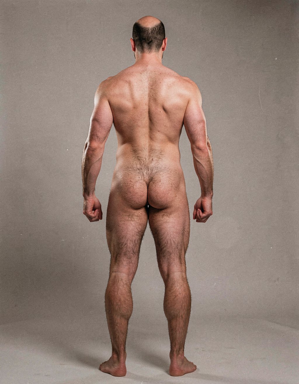 Mike Morris, a 32-year-old extremely lean Caucasian male at only 8% body fat, stands completely naked in a strict military attention pose seen from directly behind in a full posterior view. His back is perfectly straight, shoulders pulled back, arms rigid at his sides with fists clenched, legs straight and together, head held high. The shot captures him from the heels of his feet all the way up to the top of his bald head. His advanced Norwood III-IV baldness is clearly visible from behind, with a smooth tanned scalp on the crown and short dense dark brown hair only on the sides and lower back of the head. His back is highly defined and vascular: wide lats tapering dramatically to a very narrow, tight waist, thick trapezius muscles, and dense dark brown body hair lightly covering the shoulders and becoming much thicker and denser across the lower back and lumbar area, especially between the lats. His ass is tight, muscular and round with almost no fat, the glutes clearly separated. Thick dark hair continues down onto his powerful diamond-shaped calves and the backs of his slightly hairy feet. Dramatic directional studio lighting from above and slightly to the side creates strong highlights on his dry, shredded skin and deep shadows that accentuate every muscle fiber, the texture of his dense back hair, and the extreme vascularity and definition of his 8% body fat physique. Photorealistic, high-contrast, masculine 1970s Colt Studio aesthetic, grainy film texture, commanding and powerful presence. Las piernas están separadas 80 cm.  Se aprecia el surco entre glúteos  con una separación de 4 centímetros entre glúteos. Tiene el pie derecho a una distancia de ochenta centímetros del pie izquierdo