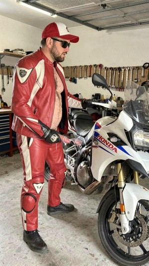 A motorcyclist in a red and white leather suit, partially open leaving his torso exposed, poses in a garage next to a white Honda motorcycle of touring or sport-touring style. He wears a red cap, sunglasses, black gloves and boots, with visible protections on knees and shoulders that reinforce the aesthetics of motorcycling gear. The motorcycle occupies the right foreground, with fairing, transparent windshield, front headlight, golden fork and visible front wheel. The environment looks like a home workshop, with white wall, raised garage door, workbench, tool cabinet and numerous wrenches hung neatly in the background, creating a scene of mechanics, maintenance and passion for motorcycles.