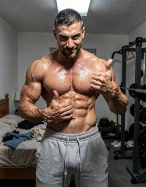 A very muscular man poses shirtless in a room that looks like a home gym, showing a defined torso, marked abs, bulky pecs, and vascular arms covered in sweat. He wears gray sweatpants with a drawstring and gives a thumbs-up gesture with both hands, looking at the camera with an intense expression and a slight smile. In the background there is an unmade bed with clothes on it, a weight rack with a barbell and plates, training equipment on the floor, and an overhead light illuminating the scene, reinforcing the atmosphere of training, bodybuilding, and fitness at home.