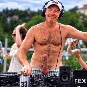 A smiling man spins music outdoors as a DJ, with large headphones, white cap and bare torso, showing his hairy chest while he handles a mixing table with turntables, cables and speakers. He wears plaid shorts pulled down enough to leave visible his erect penis sticking out above the waist, located in the center of the scene next to the sound equipment. The atmosphere looks like a party or summer outdoor event, with people out of focus in the background, green trees, clear sky and a hand holding a drink, creating a striking mix of musical performance, explicit nudity and festive atmosphere.