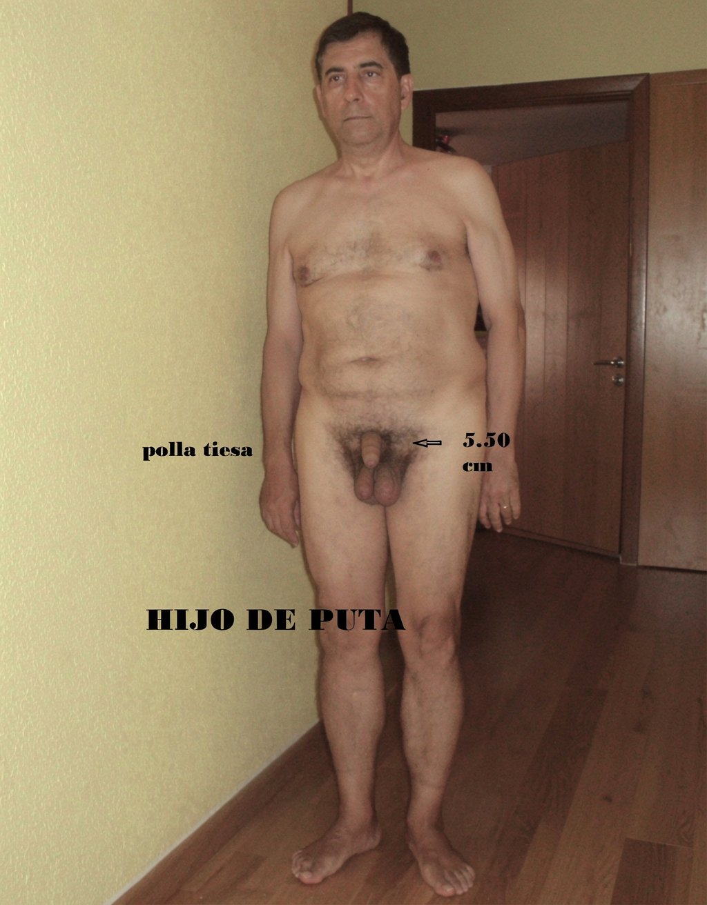 Naked man standing in an indoor hallway with a wooden floor, yellow wall, and a wooden door in the background, facing the camera with a serious expression and rigid posture, arms hanging at both sides of the body. His torso is bare, with hair on his chest and abdomen, legs slightly apart, and bare feet. The pubic area is visible, with abundant hair and the penis exposed; over the image there is black added text with an arrow pointing to the penis and the measurement “5.50 cm,” as well as the phrase “hard cock” to one side. At the bottom there is an insult written in capital letters, “SON OF A BITCH,” as an annotation superimposed on the photograph.