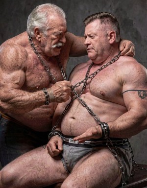 Dos hombres corpulentos y semidesnudos posan en una escena de estética BDSM sobre un fondo gris oscuro, con cadenas metálicas rodeando el cuello, el torso, las muñecas y la cintura. El hombre mayor, de pelo blanco, bigote y cuerpo musculoso, está inclinado hacia el otro con una actitud dominante, sujetando una cadena entre las manos mientras apoya un brazo sobre sus hombros. El hombre sentado, con el pecho descubierto, tatuaje en el brazo, calzoncillo gris y expresión seria, aparece atado con grilletes y cadenas cruzadas sobre el abdomen y los muslos, creando una composición intensa, física y cargada de tensión.