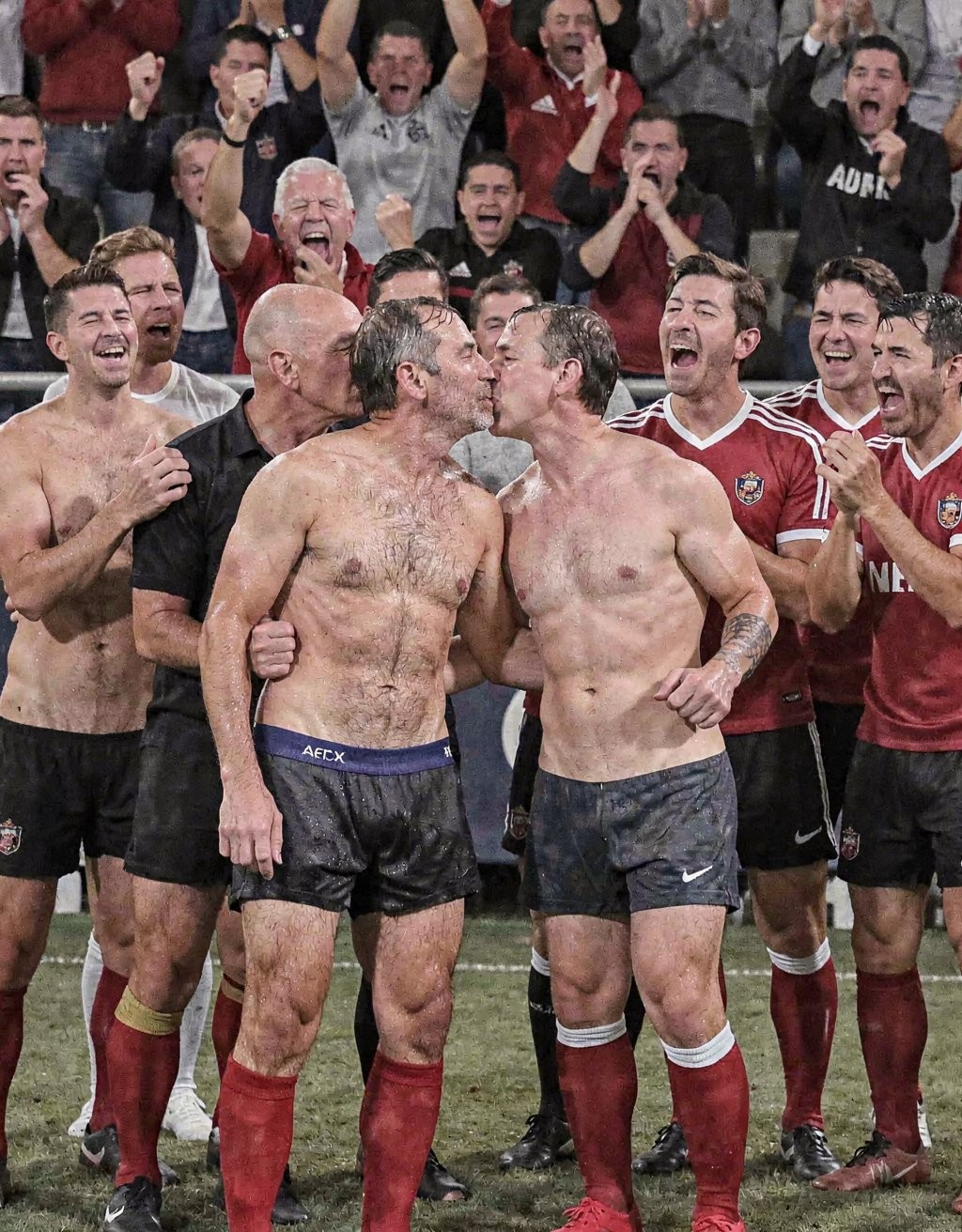 Dos hombres sin camiseta, con pantalones cortos negros embarrados y medias rojas de fútbol, se besan en el centro del campo mientras un grupo de jugadores y aficionados celebra eufóricamente a su alrededor. La escena transmite una victoria o ascenso deportivo, con rostros sonrientes, gritos, puños en alto y aplausos entre camisetas rojas del equipo, ropa mojada y barro sobre el césped. Los protagonistas, de mayor edad que la mayoría del grupo, aparecen abrazados por la cintura y rodeados de compañeros que ríen y animan, creando una atmósfera de celebración futbolística intensa, camaradería, alegría colectiva y orgullo de equipo.