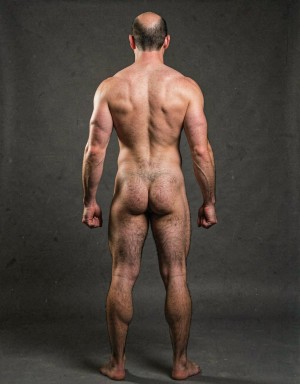 Mike Morris, a 32-year-old extremely lean Caucasian male at only 8% body fat, stands completely naked in a strict military attention pose seen from directly behind in a full posterior view. His back is perfectly straight, shoulders pulled back, arms rigid at his sides with fists clenched, legs straight and together, head held high. The shot captures him from the heels of his feet all the way up to the top of his bald head. His advanced Norwood III-IV baldness is clearly visible from behind, with a smooth tanned scalp on the crown and short dense dark brown hair only on the sides and lower back of the head. His back is highly defined and vascular: wide lats tapering dramatically to a very narrow, tight waist, thick trapezius muscles, and dense dark brown body hair lightly covering the shoulders and becoming much thicker and denser across the lower back and lumbar area, especially between the lats. His ass is tight, muscular and round with almost no fat, the glutes clearly separated. Thick dark hair continues down onto his powerful diamond-shaped calves and the backs of his slightly hairy feet. Dramatic directional studio lighting from above and slightly to the side creates strong highlights on his dry, shredded skin and deep shadows that accentuate every muscle fiber, the texture of his dense back hair, and the extreme vascularity and definition of his 8% body fat physique. Photorealistic, high-contrast, masculine 1970s Colt Studio aesthetic, grainy film texture, commanding and powerful presence. Las piernas están separadas 80 cm. La separación entre los pies es de ochenta centímetros. Se aprecia el surco entre glúteos  con una separación de glúteos de 4 cm.