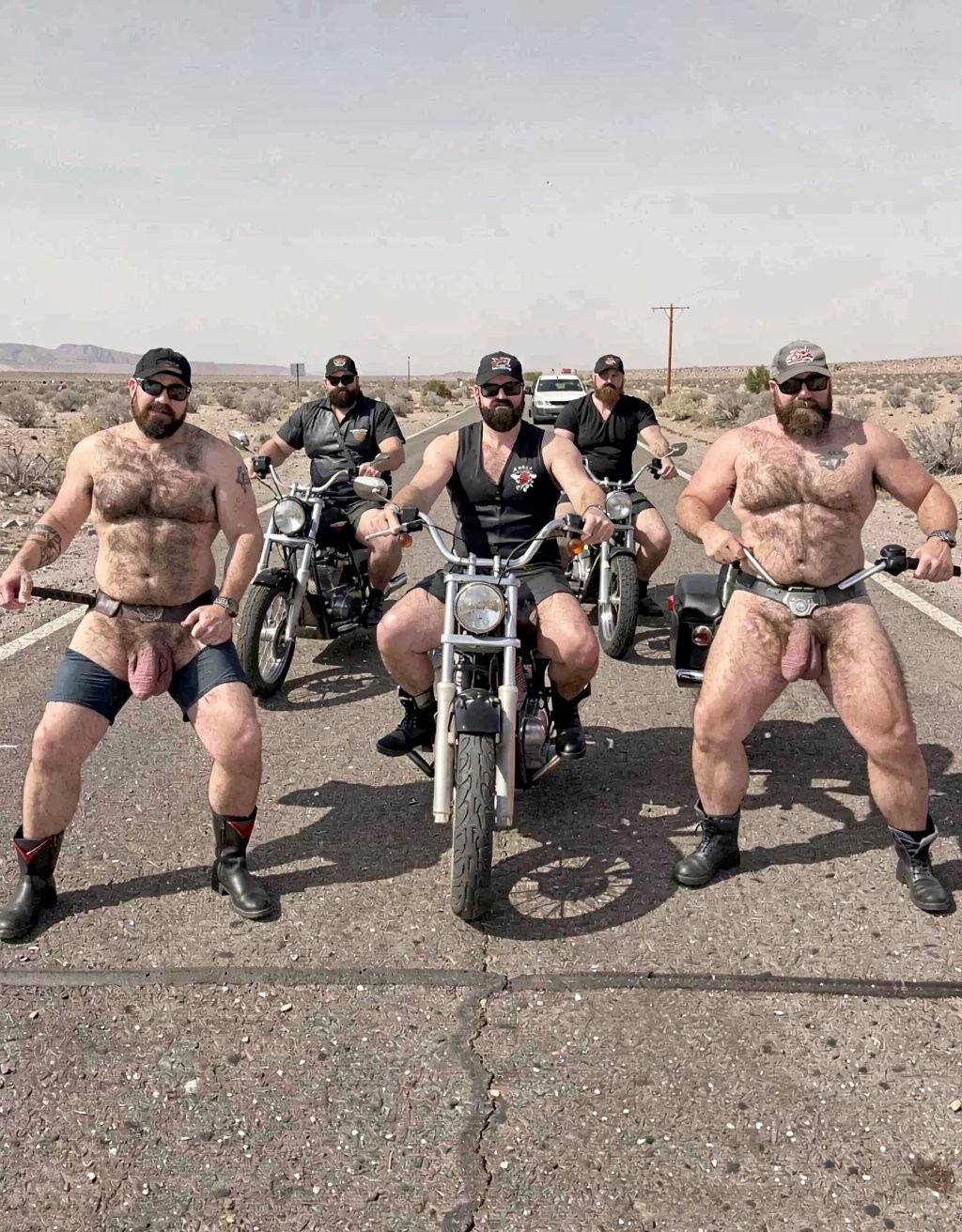 Five burly bikers pose on a desert road under a clear sky, with several cruiser-style motorcycles lined up and a dark car behind them. The two men at the ends are standing shirtless, with beards, sunglasses, black boots, and minimal leather garments in a jockstrap or loincloth style that reveal a lot of body hair and a strong leather bear aesthetic. In the center, another biker with a black vest, cap, and beard sits on the main motorcycle facing forward, flanked by two companions dressed in black on their motorcycles. The arid surroundings, cracked asphalt, road lines, and distant mountains reinforce a scene of the open road, biker masculinity, leather, desert, and defiant attitude.