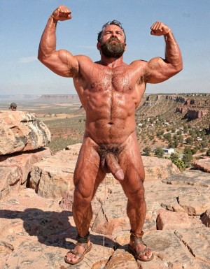 A hyper-masculine ancient Zapotec warrior named Cocijo, 26 years old, standing perfectly still under the brutal midday sun on the rocky summit of Cerro La Cantera in San Pablo Huitzo, Oaxaca, 600 AD. Completely naked except for worn leather strip sandals. Square jaw clenched, thick veiny neck, bronze skin drenched in sweat. Extremely powerful and fully flexed physique: massive square pectorals tensed to the maximum, thick dark wet chest hair matted against the hard muscle, large erect nipples. Deeply carved eight-pack abs glistening with sweat, thick black treasure trail running down to a dense dark pubic bush. Enormous arms raised and flexed hard overhead, biceps bulging like cannonballs with thick veins, triceps perfectly defined and tensed like horseshoes. Thick hairy thighs locked in full tension. His massive 35cm cock is rock-hard and throbbing violently, held tightly at the very base by Gayon Von Neumann’s strong hand, the swollen head continuously leaking thick white precum that runs down the shaft and over the fingers. A thick black leather cock ring is locked brutally tight at the root, behind his heavy balls, making the entire package look painfully swollen, darker and pulsing non-stop. Cocijo stares straight up with total obedience, every muscle in his body still tensed and displayed for inspection. Raw, dominant, extremely virile ancient masculine atmosphere, dramatic sunlight casting strong shadows on every ridge of muscle, hyper-realistic and extremely detailed.