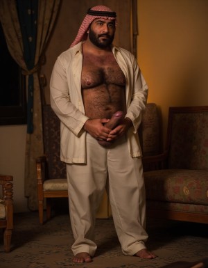 La imagen es explícita y muestra claramente el pene erecto, peludo y lleno de venas del hombre árabe. Un hombre árabe maduro de unos 50 años, con barba y bigote espesos, y un cuerpo peludo, viste un traje tradicional árabe, pero está desabrochado, mostrando su pecho velludo. Su mirada es intensa y dominante. Está de pie en una habitación lujosa con alfombras y cortinas de seda, con una iluminación cálida y sensual. La polla del maduro es gruesa de 40 cm de longitud y 8 cm de diámetro y es peluda, y está a punto de eyacular. Pone sus manos en la parte inferior del abdomen y rodea su pene para masturbarse 