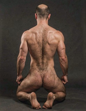 Naked man from behind, kneeling on a dark floor in a sober studio setting, with a shaved head, a broad muscular back, defined shoulders, and tense arms on both sides of the body. The light highlights the texture of the skin, the body hair running along the nape, spine, and lower back, as well as the definition of the shoulder blades, waist, and buttocks. The legs are bent beneath the body, with the soles of the feet visible and the toes pointing backward, creating a symmetrical, frontal, sculptural pose against a dark gray painterly background.