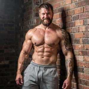 Chaser fibrado de 28 años, torso desnudo, cuerpo musculoso pero no culturista, abdominales marcados, brazos venosos, hombros anchos, mandíbula cuadrada, barba poblada cuidada, pelo corto despeinado, cejas pobladas, mirada arrogante, sonrisa de medio lado, tatuajes tribales en el brazo derecho, pantalón jogger ajustado gris marcando el paquete, cadera marcada, músculos del antebrazo tensos, en actitud dominante apoyado contra una pared de ladrillo visto, iluminación dura de contraluz, sudor ligero en el pecho.
