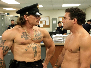 Dos hombres sin camiseta se miran cara a cara dentro de una comisaría iluminada con fluorescentes, rodeados de archivadores metálicos, tablones de anuncios, escritorios y agentes uniformados trabajando al fondo. El hombre de la izquierda lleva gorra de policía, gafas de sol, bigote, collar dorado y cinturón negro, con el torso musculoso cubierto de numerosos tatuajes en brazos, pecho y abdomen, incluida la palabra “JUSTICE” bajo el pecho; mantiene una postura desafiante con las manos en la cintura. El hombre de la derecha, también con el torso desnudo y físico atlético, le sostiene la mirada con expresión seria, creando una escena tensa y cómica de ambiente policial.
