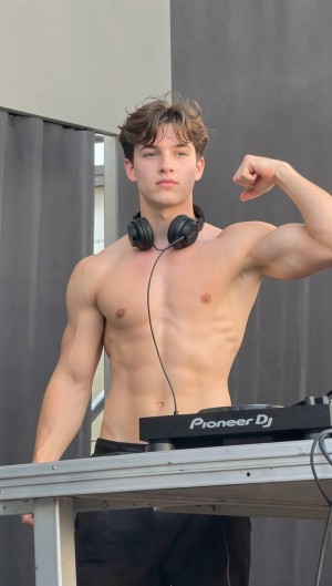 Joven DJ sin camiseta posando detrás de una mesa técnica, con unos auriculares negros alrededor del cuello y una controladora Pioneer DJ visible en primer plano. Tiene el torso musculado, abdomen marcado y levanta el brazo derecho flexionando el bíceps, mientras mira de frente con expresión seria y segura. El pelo castaño aparece despeinado de forma natural y la escena parece situada en una cabina o escenario exterior, con paneles y cortinas grises de fondo. La composición resalta el ambiente de música electrónica, actuación en directo, equipo de DJ y estética fitness.