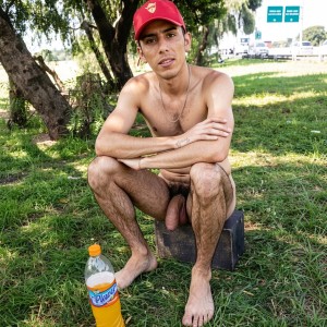 Un hombre joven está agachado sobre un bloque oscuro en una zona de césped, bajo la sombra de varios árboles, con el torso desnudo, gorra roja y una cadena fina al cuello. Mantiene los brazos cruzados sobre las rodillas y mira directamente a la cámara con expresión seria y relajada. Está descalzo, con las piernas abiertas y abundante vello corporal visible; entre los muslos se aprecia claramente el pene expuesto. En primer plano, sobre la hierba, hay una botella de bebida naranja con etiqueta visible, mientras al fondo aparecen una carretera, señales verdes y luz de día en un entorno exterior tipo parque o cuneta arbolada.