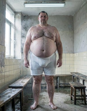 Un hombre maduro de 60 años, muy grande y gordo, posa de pie con slips blancos ajustados que marcan con claridad sus testículos muy gordos bajo la tela tensada. Se encuentra en un vestuario antiguo de azulejos crema, con bancos de madera húmeda, metal frío y un leve olor a jabón, sudor limpio y vapor. La luz principal entra lateralmente desde una ventana alta y es suave, difusa y fría, mientras un tubo fluorescente superior añade un relleno pálido que acentúa el volumen de su cuerpo y las sombras bajo la ropa interior. La escena tiene un tono íntimo, realista y fotográfico.