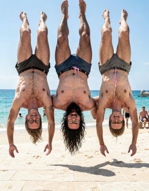 Hyperrealistic photograph captured at the exact peak of the flip in mid-air at midday on the sand of El Playazo, Vera Playa, Almería, right in front of chiringuito El Pirata. The three of you are completely naked and upside down in the middle of a full forward somersault, heads pointing straight down toward the sand, feet and legs pointing straight up to the sky, bodies perfectly inverted. Legs are spread wide open so your hairy assholes are completely exposed and centered in the frame for everyone to see, balls hanging downward by gravity, thick cocks also hanging and swinging toward your bellies.

Hipsterbarba is in the exact center: 40 years old, 1.80m, 80kg, 8% body fat, shredded dry six-pack now visible from below, dense black chest hair, trail running to your thick pubic bush, rounded pecs, wide shoulders, vascular arms, extremely long Bandholz beard hanging straight down toward the ground because of the inversion, spiral-curled mustache tips, black-rimmed glasses still on your face, high fade undercut hair, small “hipsterbarba” tattoo on your right biceps. Your hairy asshole is wide open and perfectly visible from this upside-down angle, pink ring fully exposed, heavy hairy balls dangling low, thick beautiful 19cm cock hanging down and pointing toward your chest.

To your left, Erik (38, strong hairy build, thick black beard hanging down) is also upside down with legs spread, hairy asshole exposed, thick 19cm cock and balls hanging by gravity. To your right, Ferran (36, athletic and hairy, shorter brown beard hanging down) flips in the same position, hairy asshole on full display, 17cm cock hanging freely.

All three of you have genuine laughs of pure joy on your inverted faces. Bright midday sun creates sharp highlights on every muscle, every drop of sweat flying off your bodies, and every single hair. The turquoise sea and the terrace of El Pirata are visible in the background with other naked men looking up at you, laughing and getting hard. Ultra-detailed skin texture, individual beard hairs hanging, perfect anatomical accuracy, cinematic lighting, pure joyful exhibition while fully inverted.