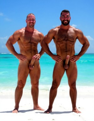 Rodrigo y Maximiliano son dos hombres culturistas de competición posando juntos en una playa de arena blanca con mar turquesa cristalino de fondo y cielo azul despejado, ambos descalzos sobre la arena, sonriendo a cámara con complicidad, postura relajada con las caderas pegadas y manos en la cintura.

Rodrigo es un hombre que está a la izquierda: caucásico de 35 años, cabeza completamente rapada y afeitada brillante por el sol, rostro cuadrado con mandíbula ancha, sonrisa amplia mostrando dientes blancos, cejas finas oscuras, ojos oscuros, piel bronceada aceitosa con brillo uniforme, cuello grueso de toro con trapecios enormes subiendo hasta las orejas, deltoides redondos y estriados como bolas de cañón con separación clara de las tres cabezas, pectorales masivos gruesos y cuadrados con estriaciones visibles en la parte inferior y surco profundo entre ambos, pezones pequeños apuntando hacia abajo por el volumen del pectoral, brazos enormes con bíceps con pico pronunciado y vena cefálica gruesa visible, tríceps con herradura marcada, antebrazos gruesos vascularizados, abdominales de seis bloques marcados con separaciones profundas, oblicuos definidos, serratos visibles, dorsales anchos que sobresalen por detrás dándole forma de V pronunciada, completamente desnudo de cintura para abajo, polla erecta de treinta y seis centímetros de longitud y ocho coma cinco centímetros de diámetro apuntando hacia arriba y ligeramente a la derecha, tronco grueso y venoso con venas retorcidas recorriéndolo desde la base hasta el glande, glande grande rosado hinchado y brillante completamente expuesto, testículos enormes gordos y pesados colgando bajos en un escroto relajado por el calor oscurecido por el bronceado, pubis recortado corto, muslos masivos con cuádriceps con forma de lágrima y separaciones marcadas, gemelos gruesos con forma de diamante, pies grandes sobre la arena.

Maximiliano es un hombre que está a la derecha: negro de piel oscura de 45 años, cabeza rapada al cero, rostro angular con pómulos altos y marcados, mandíbula cuadrada y definida, sonrisa confiada mostrando dientes blancos, ojos oscuros intensos, piel negra brillante y aceitada que resalta cada definición muscular con reflejos del sol, cuello grueso con trapecios altos, deltoides estriados enormes y redondos con venas cruzándolos, pectorales cuadrados duros y estriados con pezones oscuros pequeños, separación brutal entre ambos pectorales, brazos fibrosos masivos con bíceps vascularizados y venas gruesas serpenteando por todo el brazo, tríceps estriados con las tres cabezas visibles, antebrazos secos con cada tendón marcado, abdominales de ocho bloques profundamente tallados con piel fina pegada al músculo mostrando cada fibra, oblicuos cortados como cuchillas, serratos marcados individualmente, dorsales anchos y gruesos, completamente desnudo de cintura para abajo, polla erecta de treinta y seis centímetros de longitud y ocho coma cinco centímetros de diámetro con tronco oscuro grueso y pesado cubierto de venas hinchadas prominentes, glande enorme oscuro casi negro brillante de líquido preseminal, prepucio retraído por completo, testículos enormes gordos y llenos colgando pesados y bajos en un escroto oscuro arrugado y relajado, pubis con vello negro corto y rizado, muslos enormes fibrosos con cuádriceps estriados y femorales como cuerdas de acero, gemelos definidos y duros, pies descalzos sobre la arena blanca.

Iluminación natural directa del sol cenital con reflejos en la piel aceitada de Rodrigo y Maximiliano, sombras cortas en la arena, alta resolución fotográfica hiperrealista, textura de piel con poros visibles, gotas de sudor y aceite, calidad de fotografía profesional editorial deportiva, plano frontal de cuerpo entero.



No distorsiones y NO DEFORMACIONES.