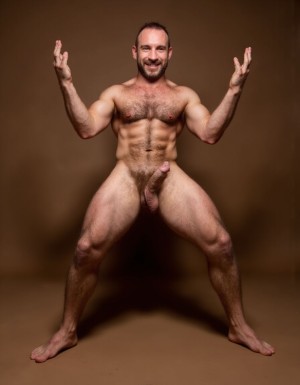 hombre culturista de 35 años posando desnudo de cuerpo completo, sonriente, tensa los bíceps, tiene un pene de 40 cm de longitud y 7 cm de diámetro, grandes dorsales, piernas, cintura estrecha, un poco calvo, grandes pezones rosas. Tiene un pene de 40 cm de longitud y 7 cm de diámetro. Tiene mucho vello en pecho y brazos y piernas. Tiene testículos de 10 kg de peso y están colgando. Tiene pies con sus uñas rojas. Fondo de color marrón. Está agachado con las piernas abiertas. 