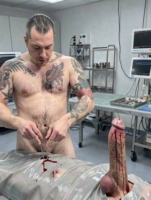 Un hombre desnudo y tatuado aparece en un entorno clínico o quirúrgico, de pie junto a una camilla cubierta con plástico transparente manchado de sangre, mientras manipula unas pinzas o instrumental médico sobre una pequeña herida o zona ensangrentada a la altura de la pelvis. Tiene el torso descubierto, varios tatuajes en pecho y brazos, y la cabeza inclinada con gesto concentrado. A la derecha, sobre la camilla, destaca un pene erecto de aspecto hiperrealista o protésico, cubierto de sangre que escurre desde la punta y por el tronco hasta los testículos. Al fondo hay monitores médicos, cables, bandejas metálicas, carros auxiliares e instrumental, reforzando la sensación de procedimiento médico extremo, modificación corporal o escena de cirugía experimental.