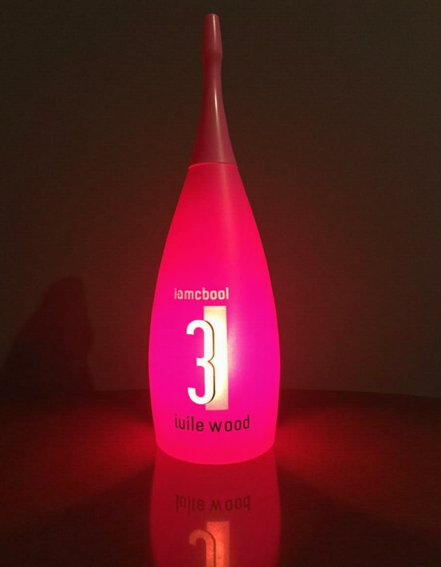 Decorative lamp with an elongated bottle or diffuser shape, illuminated from within by an intense fuchsia-pink glow that tints the glossy surface it rests on. The translucent body narrows toward a thin neck capped with a grayish tip, creating an elegant vertical silhouette against a dark background. On the front appears the text “iambcool,” a large white number “3,” and the inscription “huile wood” in small letters, all integrated into the luminous design. The table partially reflects the figure and the reddish glow, reinforcing the nocturnal, modern, and minimalist atmosphere.
