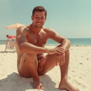 A naked, smiling man poses seated on the sand of a sunny beach, with his muscular torso exposed, his legs bent, and his arms resting relaxed on his knees. His penis is visible between his legs, in a scene of natural nudity with no explicit sexual gesture. In the background, the blue sea, the horizon line, a striped beach umbrella, and a person next to a chair can be seen, all under warm summer light that reinforces the vacation-like, calm, and bright atmosphere.