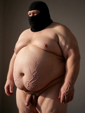 Hombre desnudo de complexión muy corpulenta posando de pie contra un fondo gris oscuro, con un pasamontañas negro que cubre casi toda la cabeza y deja visibles solo los ojos. Su torso está descubierto, con pecho amplio, barriga prominente y ombligo marcado, además de unas formas ramificadas en relieve sobre la piel del abdomen que recuerdan a venas, cicatrices o raíces. La iluminación lateral resalta los pliegues de la piel, los brazos robustos y la textura corporal. En la parte inferior aparece el pene flácido y el escroto visibles entre los muslos, mientras sus manos cuelgan relajadas a ambos lados del cuerpo, creando una escena cruda, frontal y de estudio.