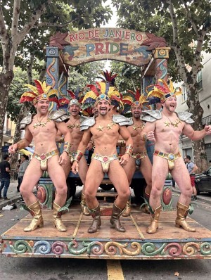 Five smiling men dance on a festive float decorated with tropical motifs and bright colors, beneath a large arch that says “Rio de Janeiro Pride.” They wear carnival costumes inspired by Brazilian aesthetics, with multicolored feather headdresses, silver shoulder pads, bracelets, ornate belts, golden boots, and beige briefs with gold and turquoise details that leave the torso and legs exposed. The scene conveys the energy of a pride parade, LGBTQ+ celebration, music, and street performance, with trees, spectators, and urban buildings in the background, while the platform features painted decorations in pastel tones and wavy shapes.