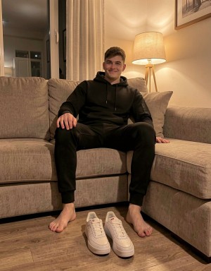 Un joven sonriente está sentado relajadamente en un sofá rinconera beige, con las piernas separadas y los pies descalzos apoyados sobre el suelo de madera. Viste un conjunto deportivo negro compuesto por sudadera con capucha y pantalón de chándal, creando un estilo informal y cómodo de estar en casa. Delante de él, perfectamente colocadas, hay unas zapatillas blancas limpias con cordones, como preparadas para ponérselas. La escena tiene una iluminación cálida y acogedora gracias a una lámpara de pie encendida junto al sofá, con cortinas claras, una ventana al fondo y un cuadro en la pared que refuerzan el ambiente doméstico y tranquilo de salón moderno.