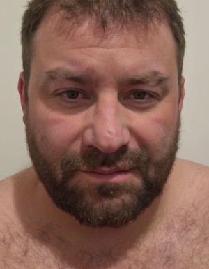 Close-up of a man with his face centered, short brown hair somewhat disheveled and a thick beard well defined around the jaw and chin. He looks directly at the camera with a relaxed expression and a slight smile, light eyes slightly narrowed, thick eyebrows and somewhat flushed cheeks. His bare shoulders and part of his chest with hair appear, suggesting he is shirtless. The background is plain and light, without decorative elements, with soft indoor lighting that highlights facial features, skin texture, beard and hair in an intimate close-up portrait composition.