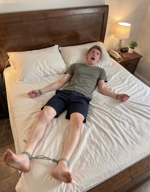 Un hombre joven aparece tumbado boca arriba sobre una cama grande con sábanas blancas arrugadas y un cabecero alto de madera oscura, vestido con camiseta gris y pantalón corto azul marino. Tiene los brazos extendidos a los lados y las piernas estiradas, con los tobillos atados con una cuerda gris cerca del extremo de la cama, lo que crea una escena de inmovilización o juego de ataduras. Su expresión es de sorpresa o sobresalto, con la boca abierta y la mirada dirigida hacia arriba. A la derecha hay una mesilla de noche con una lámpara encendida, un teléfono, pequeños objetos y una planta decorativa, aportando un ambiente de dormitorio doméstico cálido y ordenado.