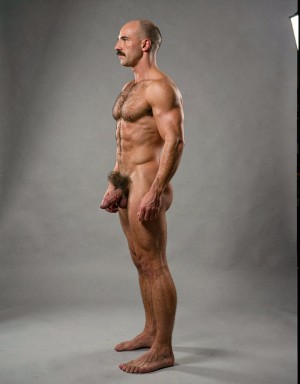 Mike Morris, a 32-year-old extremely lean Caucasian male with only 8% body fat, stands completely naked in a rigid military attention pose captured in a full-body exact side profile at 90 degrees from his right side. The shot runs uninterrupted from the tips of his size 44 slightly hairy feet all the way up to the top of his bald head, showing his entire body without any cropping whatsoever. His physique is dry, highly vascular and powerfully defined: massive square pectorals seen in perfect side silhouette, dense dark brown chest hair forming a thick T-pattern that continues across his shoulders and back, sharply carved six-pack abs in clear profile, narrow waist, wide flaring lats, powerful thighs and diamond-shaped calves covered in the same dark hair. Dramatic directional studio lighting falls from high and slightly in front of him, creating strong rim lighting that traces every sharp edge of his muscular outline, accentuating the texture of his dense dark body hair on chest, shoulders, lower back, ass and legs, while producing deep shadows that highlight every muscle separation, vein and the extreme dryness of his 8% body fat. His advanced Norwood III-IV bald scalp is clearly visible under the light, with very short dense dark brown hair only on the sides and back of his head. His square, angular face is shown in pure right profile: strong jawline, prominent chin with slight central dimple, thick well-groomed dark handlebar mustache, heavy brow and intense deep-set brown eye staring straight forward with a serious, commanding expression. His penis is fully and clearly visible in perfect side view, completely natural and anatomically correct: thick, 24 cm long when fully erect, with a natural slight upward curve, prominent dorsal veins running along the shaft, long foreskin that partially covers the pink-red glans, and noticeably lighter skin tone than the rest of his bronzed body. Below it hang his large, low-hanging testicles with wrinkled darker skin and an extremely dense, curly dark brown pubic bush that climbs almost to his navel and surrounds the base of his cock and balls generously. Every detail from his veined hands and forearms to his muscular ass, hamstrings, diamond calves and hairy toes is perfectly proportioned and free of any deformation. Photorealistic, high-contrast, masculine 1970s Colt Studio aesthetic with subtle grainy film texture.