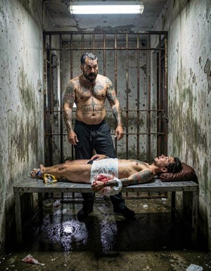 A heavyset man, shirtless and covered in tattoos, stands in a narrow, deteriorated cell, his torso stained and a hard, almost defiant expression on his face under cold fluorescent light. In front of him, another tattooed man lies motionless on a metal table, dressed only in white underwear, his head turned on a makeshift pillow and several visible bloodstains near his waist and side. The setting looks like an abandoned prison or torture chamber, with rusted bars in the background, damp walls full of mold, peeling paint, a flooded floor and dirty scattered debris, creating a violent, oppressive and raw scene of captivity, assault and prison brutality.