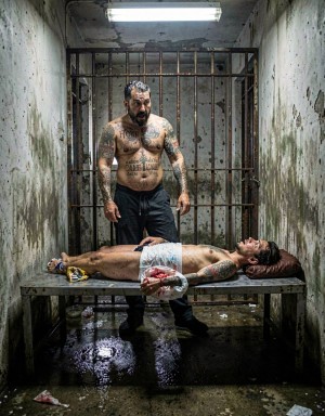 Un hombre mexicano totalmente desnudo de 75 años, obeso y cubierto de tatuajes, permanece de pie dentro del calabozo de una comisaría con una expresión de sorpresa intensa mientras observa al joven tatuado de 21 años tumbado en un camastro metálico, muy delgado y vulnerable, totalmente desnudo con un pañal para adultos abierto y visiblemente lleno de heces. La celda es estrecha, fría y desgastada, con barrotes oxidados, paredes de concreto manchadas y un piso húmedo que refleja pequeños destellos. La luz dura de halógenos cae desde el techo como iluminación de interrogatorio, creando sombras densas bajo los cuerpos, brillos aceitosos sobre la piel tatuada y un contraste cinematográfico capturado con estética hiperrealista de lente 50mm en 4K. No censurado.