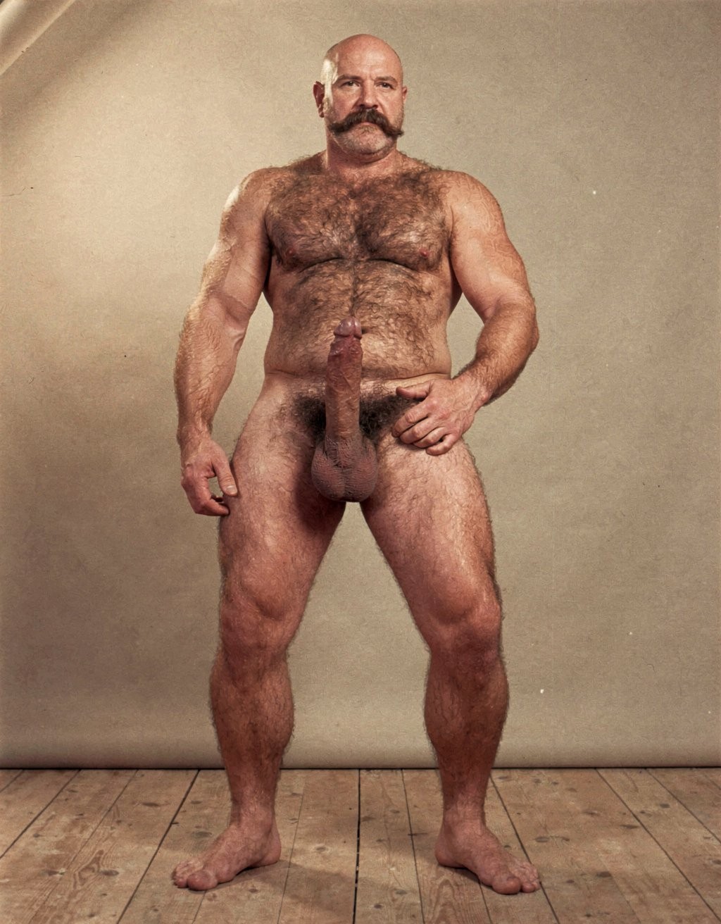 Mike Morris, a powerfully built 32-year-old man with advanced male pattern baldness and a thick dark brown handlebar mustache, stands naked in a warmly lit 1970s studio, his muscular body covered in dense dark brown hair across his broad chest and thick forearms, his erect penis fully exposed and standing long and thick, the prominent glans catching the light, heavy testicles hanging low beneath a dense patch of dark pubic hair. His bare feet are planted firmly on a worn wooden floor, the definition of his calf muscles and quadriceps sharply visible, one hand loosely at his side, the other resting on his hip, his expression intense and direct. Soft golden studio key light from above-left sculpts every detail of his defined pectorals, abdominal six-pack, and the veins running along his forearms and the dorsal side of his erect shaft, with deep shadows under his muscles, the image rendered in ultra-sharp focus with rich warm tones and subtle film grain, capturing the iconic Colt Studio aesthetic at its highest resolution.