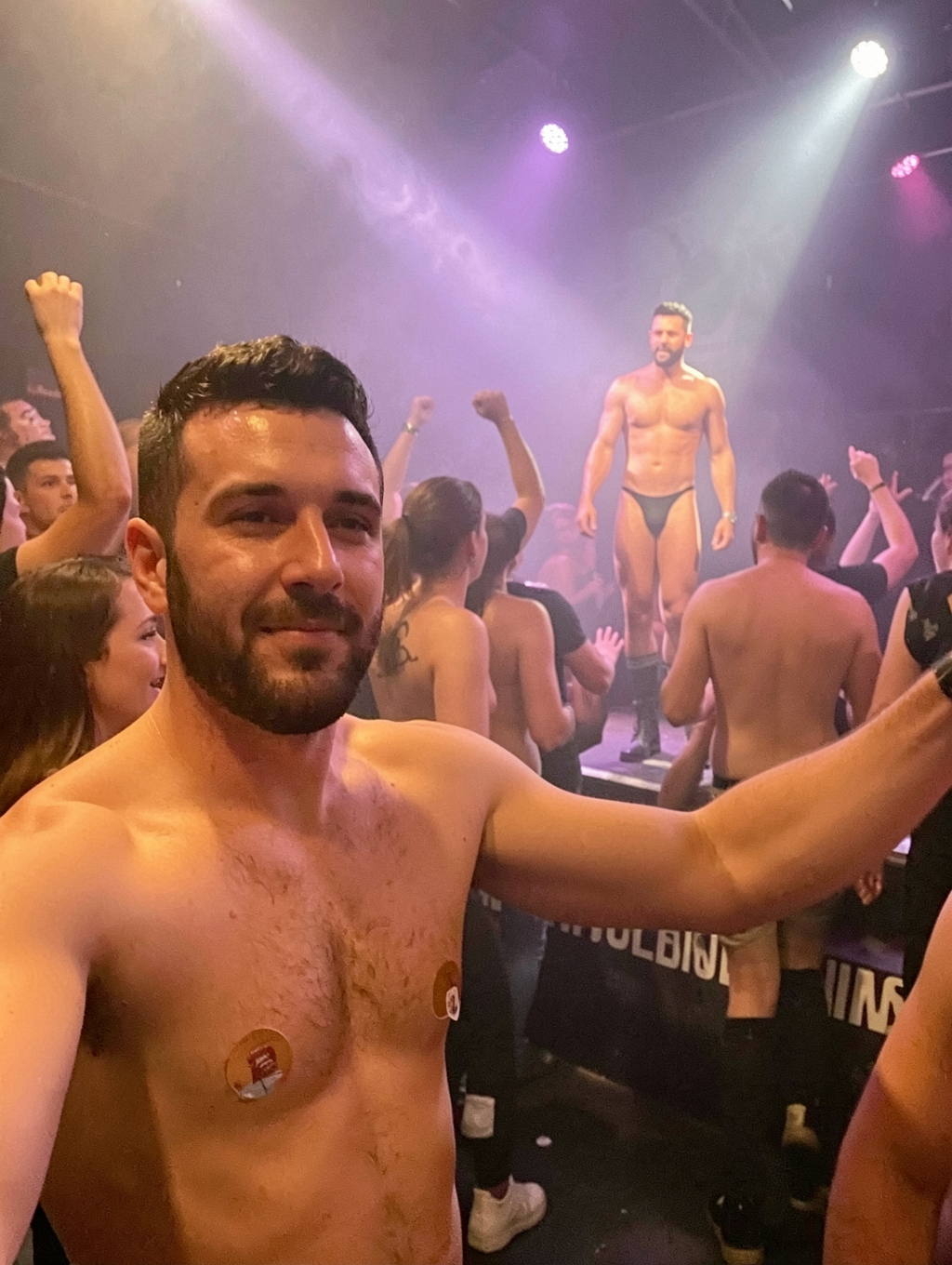 A shirtless man takes a selfie in close-up inside a club or night party, with his torso sweaty, trimmed beard and a circular sticker on his chest, while smiling slightly and raising his arm to frame the scene. In the background, among purple and white lights and disco smoke, a muscular dancer poses on an elevated platform wearing black briefs or a thong, illuminated by stage spotlights. Around him is a crowd of men, many also shirtless, dancing, raising their arms and looking toward the show, creating an atmosphere of party, music, dancing, pride and gay disco energy.