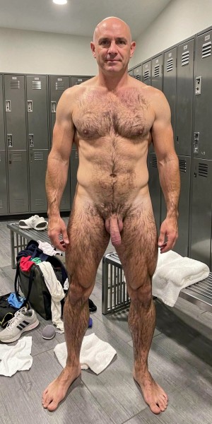 Naked man standing in a locker room, looking directly at the camera with frontal posture and arms relaxed at his sides. He has a shaved head, muscular and athletic build, torso and legs with abundant body hair, broad chest, marked abdomen and visible penis between his thighs. The environment consists of gray metal lockers, locker room benches, a folded white towel and messy sportswear next to some sneakers on the floor. The artificial ceiling lighting highlights the skin, musculature and the functional atmosphere of a gym or shower area.