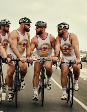 Cinco chicos de mediana edad, cuerpos grandes musculosos con barba corta, todos con traje de triatlón blancos marcando enormes bultos  en la bicicleta, pertenecen a un equipo de triatlón gay y llevan distintivos de este equipo, casco y gafas iguales. Se miran fijamente. 