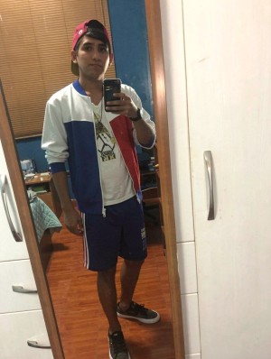 Un joven posa frente a un espejo de cuerpo entero haciéndose un selfi con el móvil en una habitación. Lleva una gorra roja colocada hacia atrás, chaqueta deportiva blanca con paneles azul y rojo, camiseta blanca con estampado gráfico, pantalón corto azul y zapatillas negras. Su postura es relajada, con una mano sujetando el teléfono y la otra junto al cuerpo, mientras mira hacia el reflejo. El entorno muestra un dormitorio con suelo de madera, armario blanco con tiradores metálicos, una cama parcialmente visible, una mesa con objetos y una persiana bajada al fondo, creando una escena cotidiana e informal de moda urbana y estilo casual.