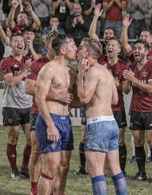 Dos jugadores de rugby sin camiseta se besan en el centro del campo durante una celebración, con el cuerpo sudado y embarrado tras el partido; uno lleva pantalón corto azul oscuro con el escudo del equipo y el otro un short azul claro, medias altas y una mano apoyada cerca de la barbilla de su compañero. Alrededor, varios compañeros con camisetas granates y pantalones negros aplauden, gritan y levantan los brazos con euforia, mientras el público en las gradas también celebra y algunos fotografían el momento. La escena transmite alegría, orgullo, compañerismo deportivo y emoción intensa en un ambiente de estadio, con césped mojado, uniformes manchados y una atmósfera de victoria colectiva.