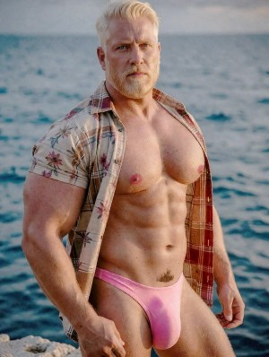 Muscular man with short blond hair and trimmed beard posing by the sea, with a serious expression and direct gaze at the camera. He wears an open short-sleeved shirt with floral and checkered print, revealing his defined torso, pectorals, marked abs, and strong arms. He wears a pink thong swimsuit with a very high cut, snug at the crotch, which highlights the pubic area and legs. The background shows blue water with small waves and natural outdoor light, creating a beach or coastal scene with a summery, athletic, and provocative aesthetic.