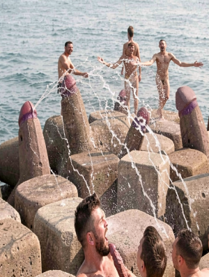 Several naked men play and pose on large concrete blocks of a breakwater by the sea, surrounded by water splashing in jets that cross the scene. Some structures are painted and sculpted in the shape of giant penises, with pink and purple glans, creating a provocative and festive atmosphere. In the background, three men smile and raise their arms over the artificial rocks, while in the foreground other participants appear partially visible among the splashes. Daylight, the waves and the coastal setting reinforce the feeling of an outdoor celebration, with male nudity, sexual humor and a striking composition between phallic sculpture, beach, sea and breakwater.