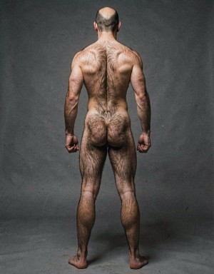Un hombre desnudo aparece de espaldas en un estudio con fondo gris oscuro, de pie y con las piernas ligeramente separadas, mostrando una complexión musculosa y muy definida. La espalda ancha, los hombros marcados, los brazos tensos y los puños cerrados transmiten una postura firme y atlética, mientras el vello corporal recorre la nuca, la columna, los glúteos y las piernas. La cabeza rapada en la coronilla y la iluminación lateral resaltan la textura de la piel, los músculos dorsales, la cintura estrecha, los glúteos expuestos y las pantorrillas fuertes, creando una composición sobria de desnudo masculino artístico.