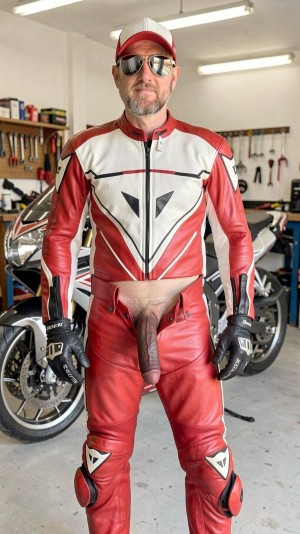 Un hombre con barba, gafas de sol y gorra roja posa de pie en un garaje o taller, vestido con un mono de motociclismo de cuero rojo, blanco y negro con protecciones y guantes negros, mientras varias motocicletas y herramientas colgadas aparecen al fondo bajo luces fluorescentes. El traje está abierto en la zona frontal de la entrepierna, dejando al descubierto el pubis y el pene flácido, lo que contrasta con el aspecto técnico y deportivo del equipamiento de piloto. La escena tiene un ambiente de garaje mecánico, con suelo de cemento, bancos de trabajo y motos aparcadas, y el hombre mira directamente a la cámara con una postura firme y segura.