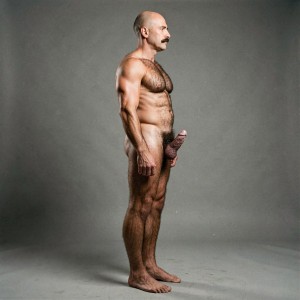 Mike Morris, a 32-year-old extremely lean and dry Caucasian male with only 8% body fat, stands completely naked in a rigid military attention pose captured in a full-body exact side profile at 90 degrees from his right side. The shot runs uninterrupted from the tips of his size 44 slightly hairy feet all the way up to the top of his bald head, showing his entire body without any cropping whatsoever. His physique is exceptionally defined and vascular: massive square pectorals seen in perfect side silhouette covered in dense dark brown chest hair forming a thick T-pattern that spreads across his shoulders and lower back, sharply carved six-pack abs in clear profile, narrow waist, wide flaring lats, powerful thighs and diamond-shaped calves all covered in the same thick dark hair. Dramatic directional studio lighting falls from high and slightly in front of him, creating strong rim lighting that traces every sharp muscular edge, accentuating the texture of his dense dark body hair on chest, back, ass and legs while producing deep shadows that highlight every vein, muscle separation and the extreme dryness of his 8% body fat. His advanced Norwood III-IV bald scalp shines under the light, with very short dense dark brown hair only on the sides and back. His square, angular face is shown in pure right profile with strong jawline, prominent chin with slight central dimple, thick well-groomed dark handlebar mustache, heavy brow and intense deep-set brown eye staring straight forward with a serious, commanding expression. His penis is fully and clearly visible in perfect side view, completely natural and anatomically correct: thick, 24 cm long when fully erect, with a natural slight upward curve, prominent dorsal veins, long foreskin that partially covers the pink-red glans, and noticeably lighter skin tone contrasting against his bronzed body. Below it hang his large, low-hanging testicles with wrinkled darker skin surrounded by an extremely dense, curly dark brown pubic bush that climbs almost to his navel. Every anatomical detail from his veined hands and forearms to his muscular ass, hamstrings, diamond calves and hairy toes is perfectly proportioned and realistic. Photorealistic, high-contrast, masculine 1970s Colt Studio aesthetic with subtle grainy film texture.